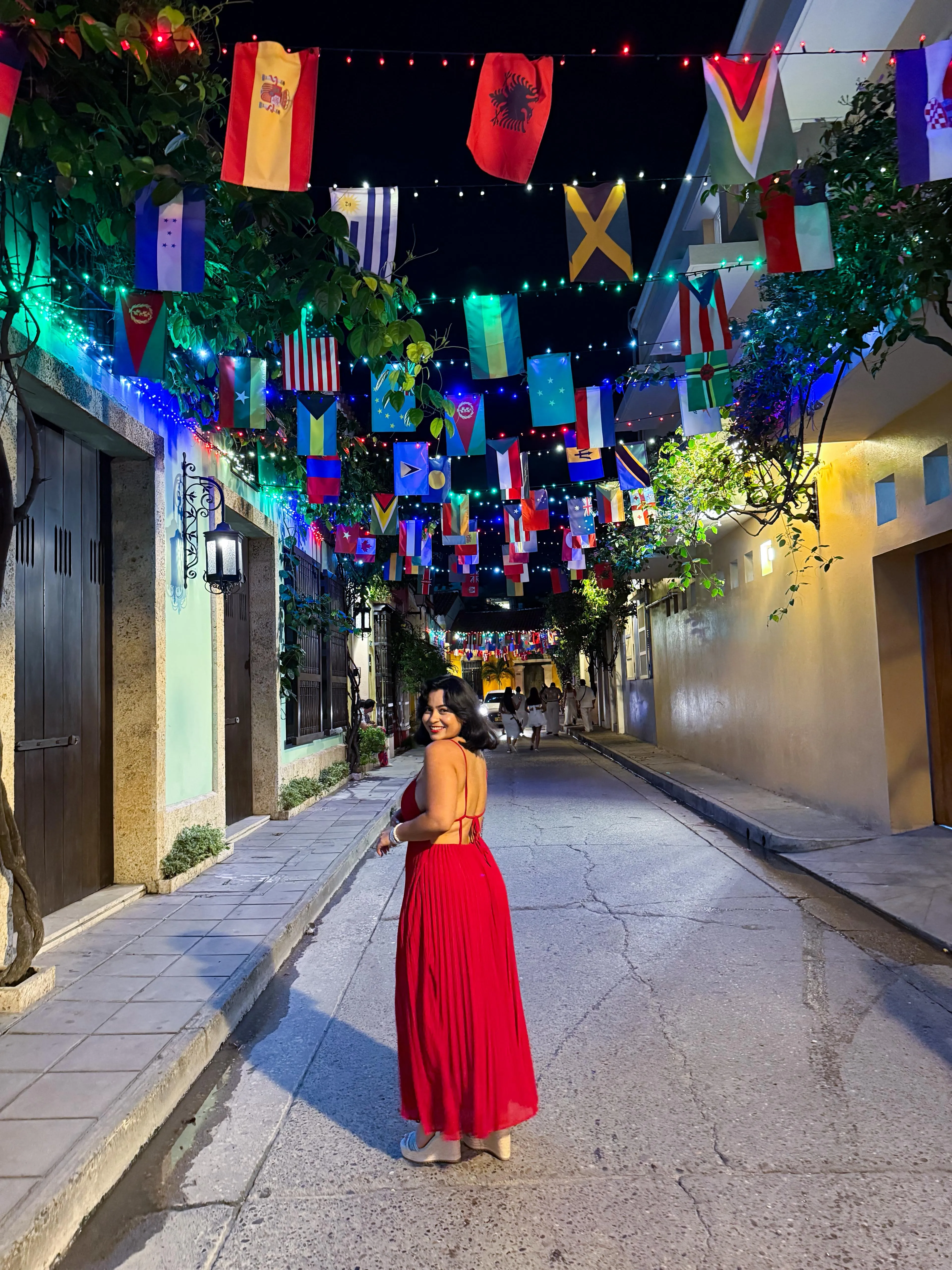 A woman in a red dress looks back over her shoulder in a flag-lit Cartagena alley at night