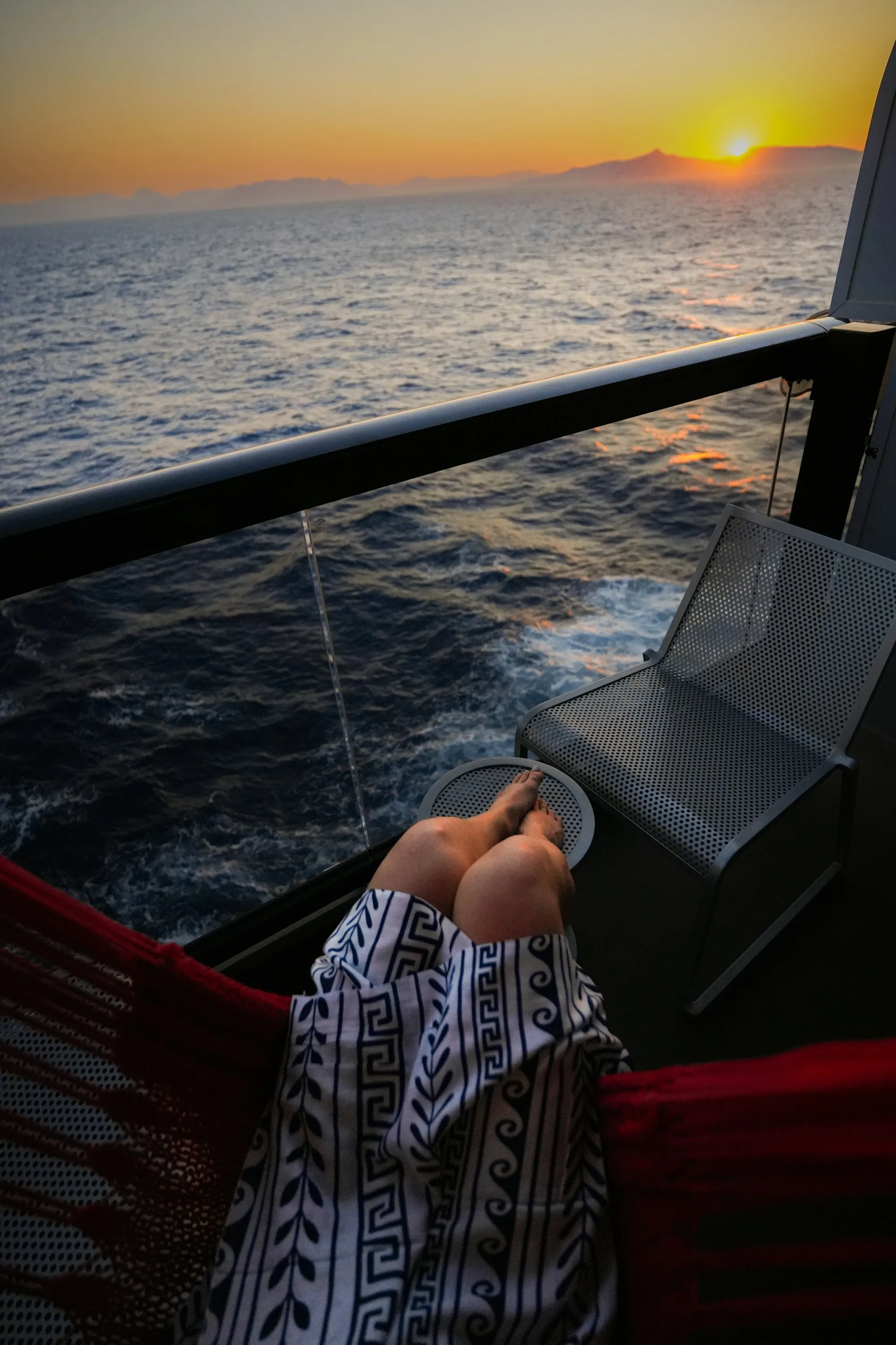 A person's silhouette resting in a hammock facing the ocean at sunset